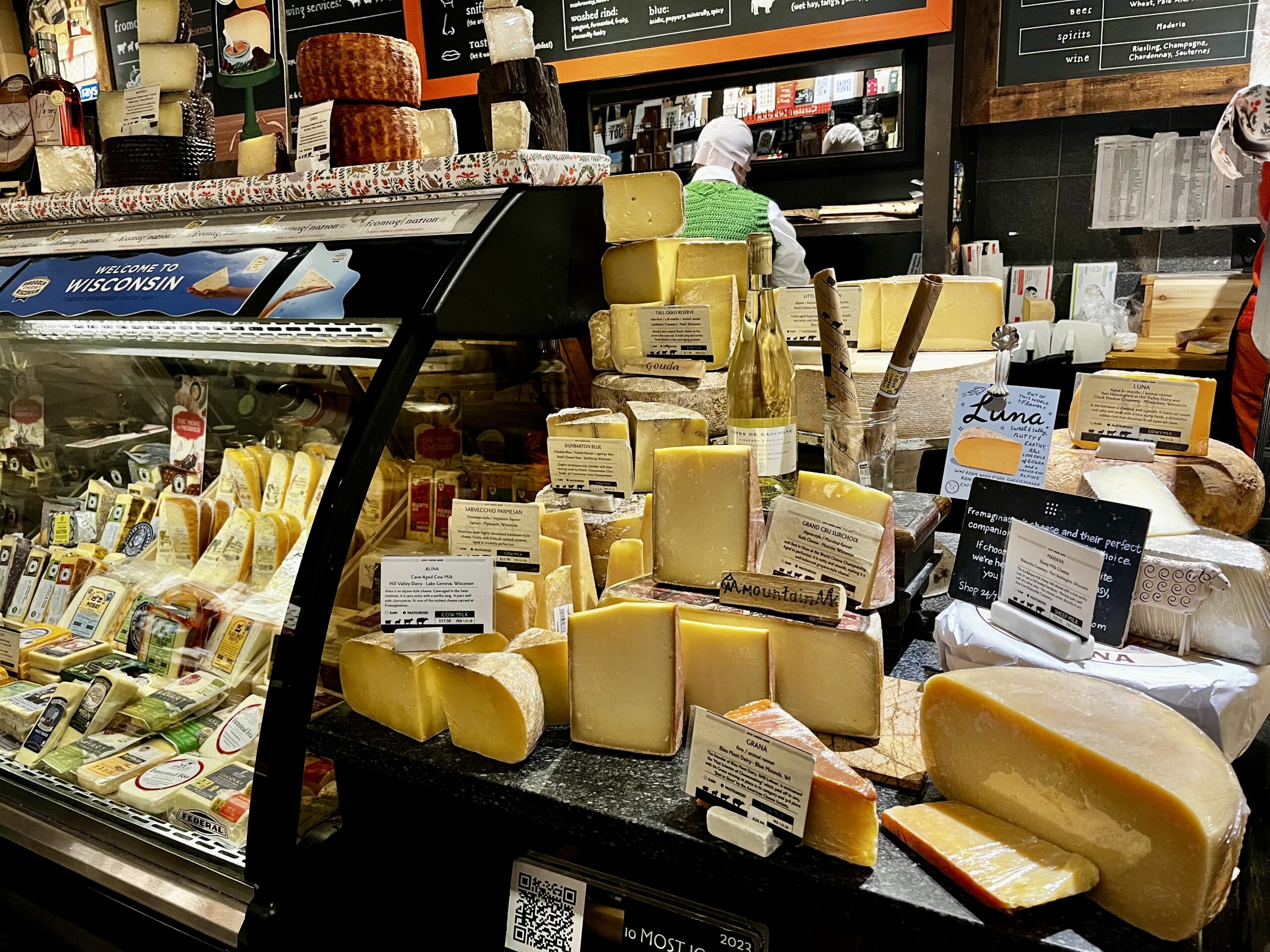 Interior of an artisan cheese shop with a black granite counter displaying various Wisconsin cheese wheels and blocks, including wrapped and unwrapped varieties, with a refrigerated case of additional cheeses on the left and a "Welcome to Wisconsin" sign visible.