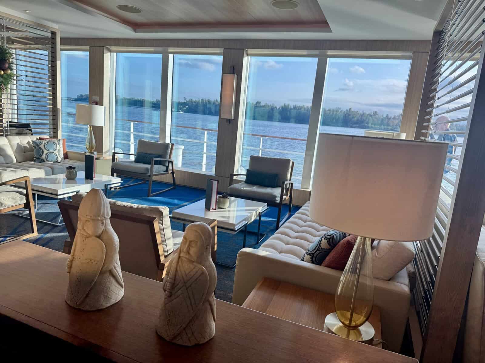 Two Nordic figurines in the foreground, white sofas and gray chairs adorn a living room, with a view of the river through large windows.