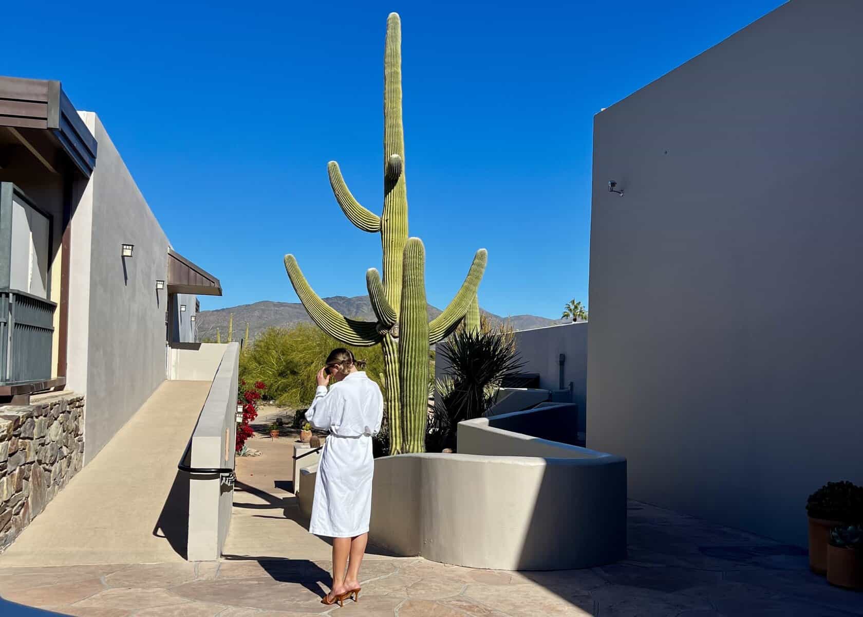  Woman in a white robe, heels and sunglasses stands next to a cactus.