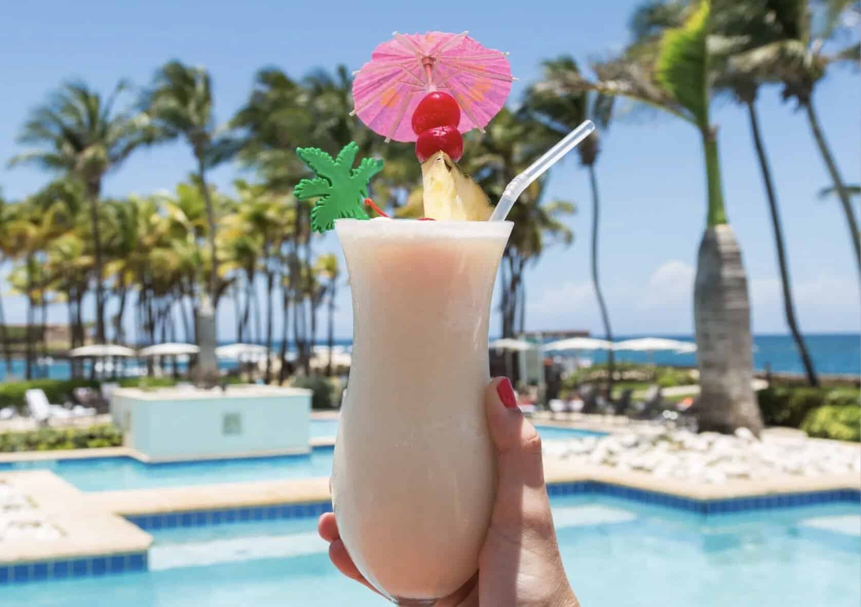 Tall glass with creamy drink topped with a wedge of pineapple, a cherry and a pink umbrella with a pool in the background