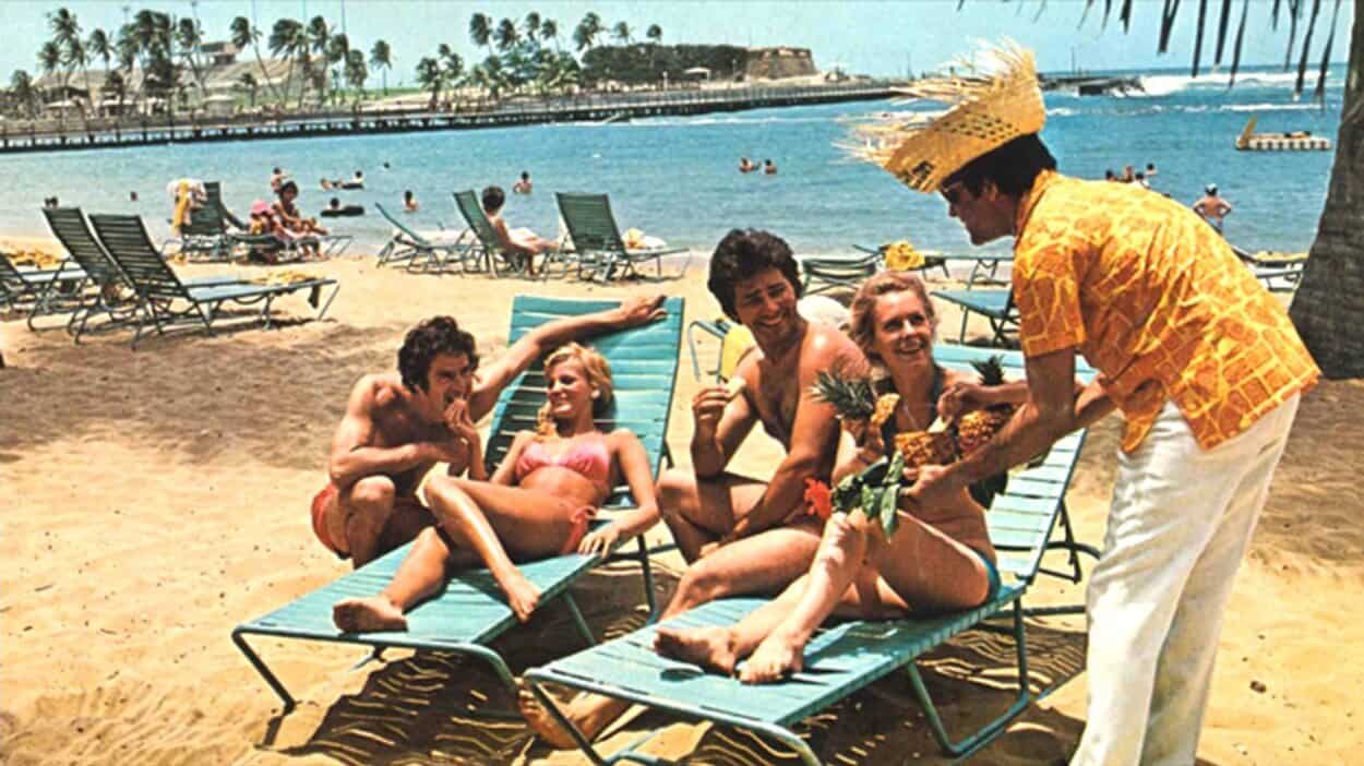  Beach waiter serves Piña Coladas in pineapples to four guests in blue lounge chairs