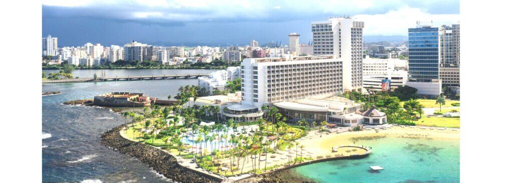 Aerial view of white hotel with pool and palm trees bordering the ocean