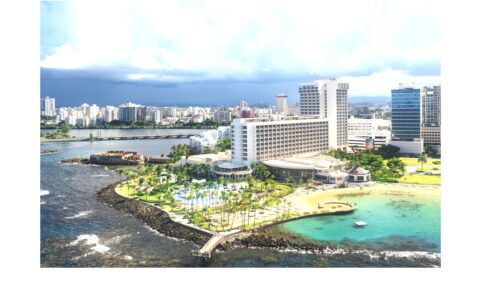 Aerial view of white hotel with pool and palm trees bordering the ocean