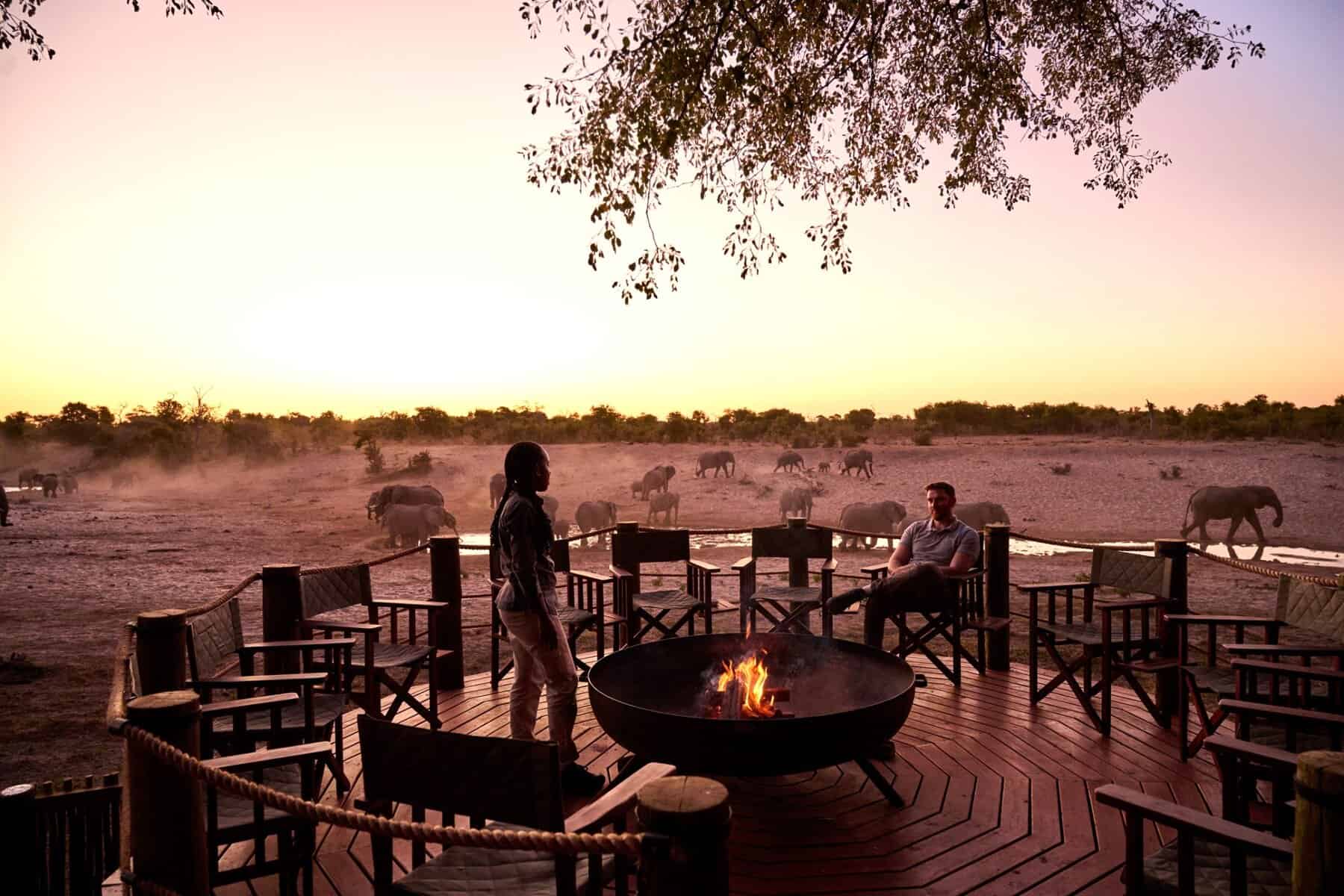 Sunset fire pit deck with wildlife at waterhole and dramatic sky at Savute Safari Lodge.