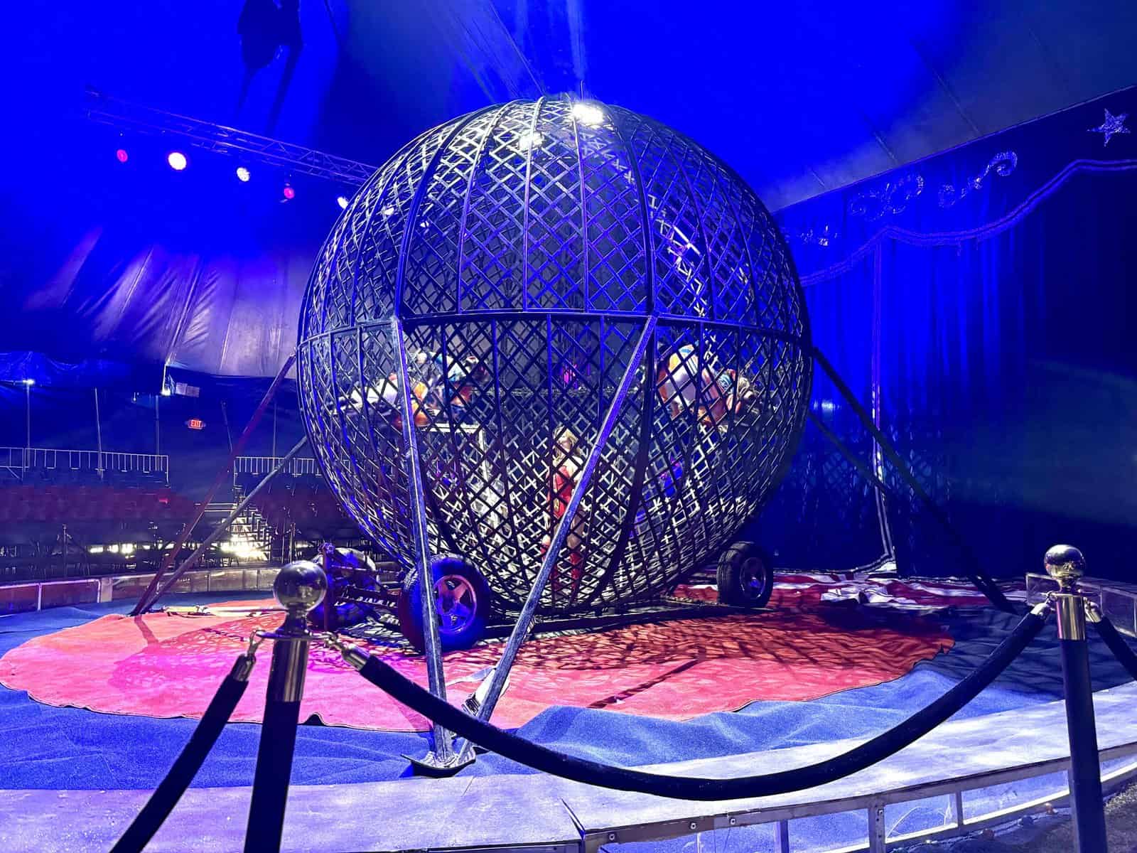 Two men ride motorcycles around a standing female inside a metal sphere.