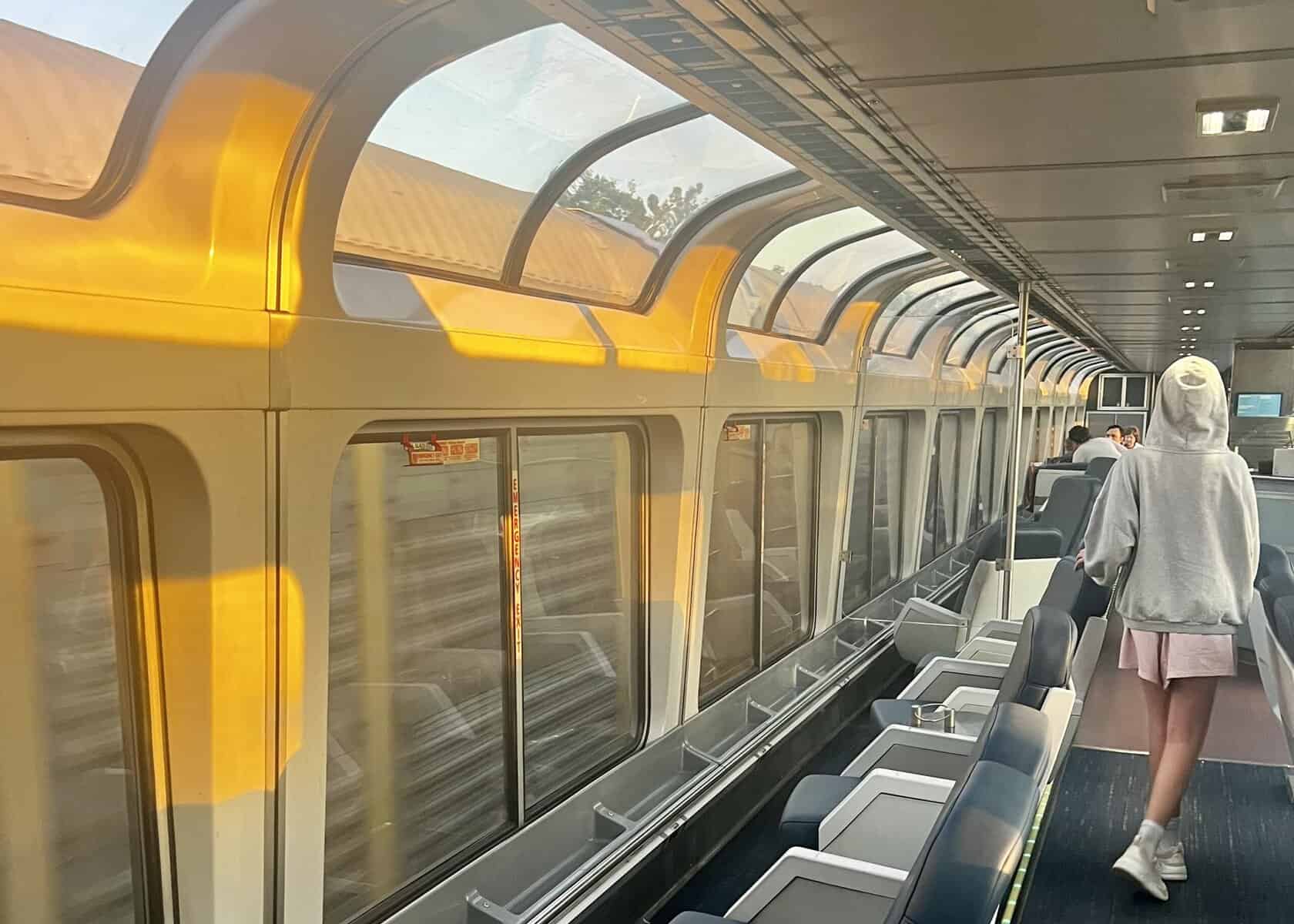 Girl in pink shorts walks through train car with wall and ceiling windows