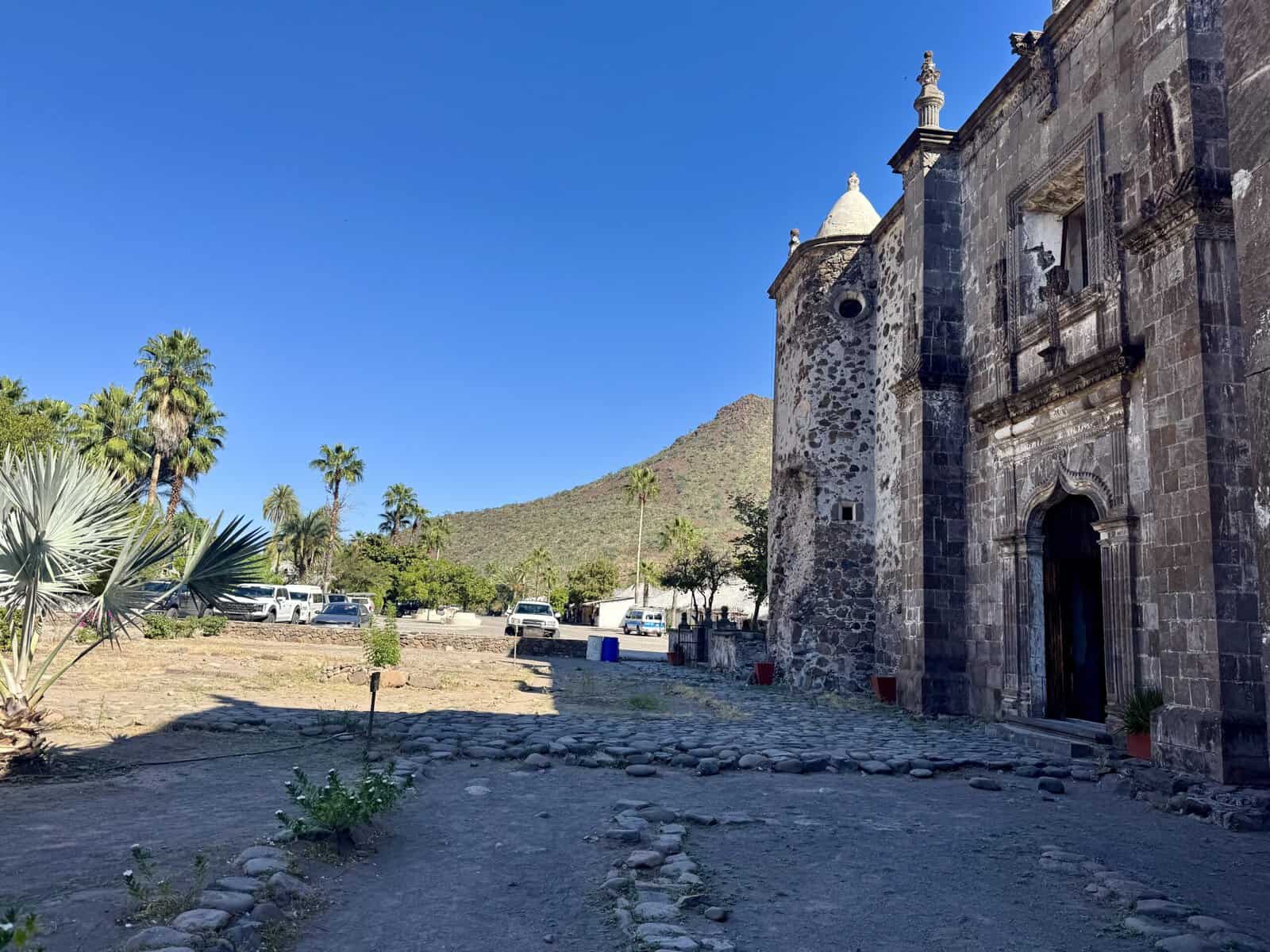 Stone mission building surrounded by palm trees and mountains.