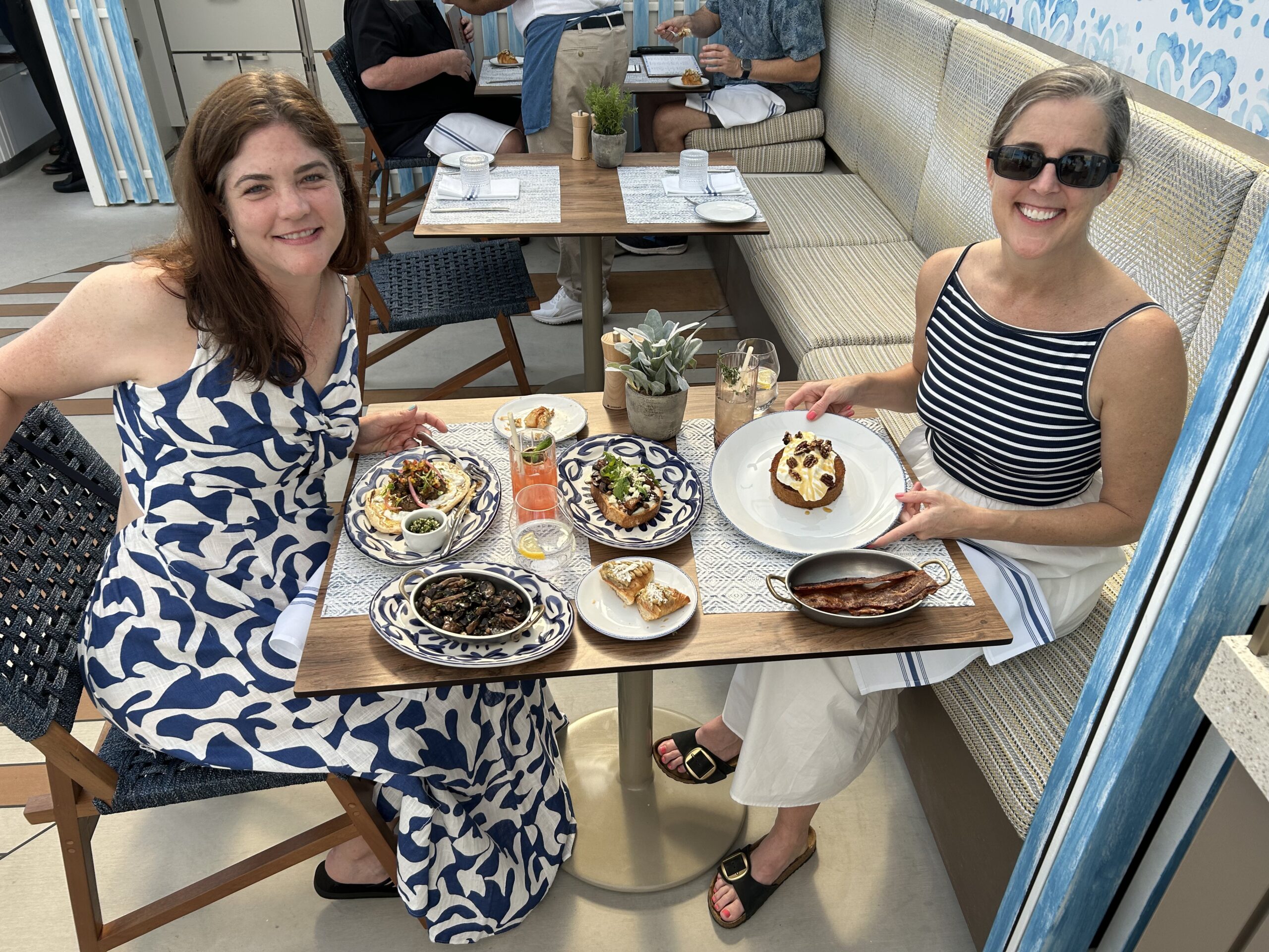  Two women smiling over a table of food in a bright blue-and-white Mediterranean-style restaurant.