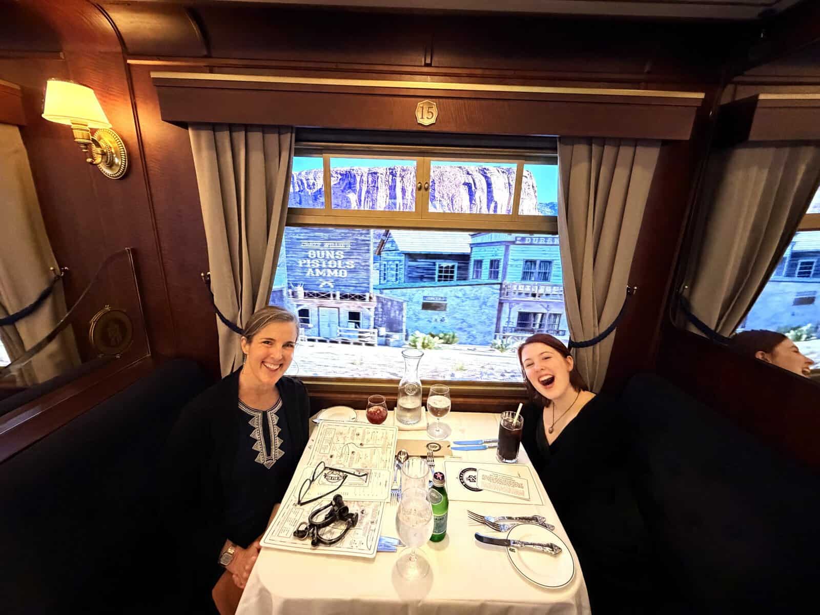 Two women laughing in a period train car dining room with LED projection of a Western frontier town visible through the window.