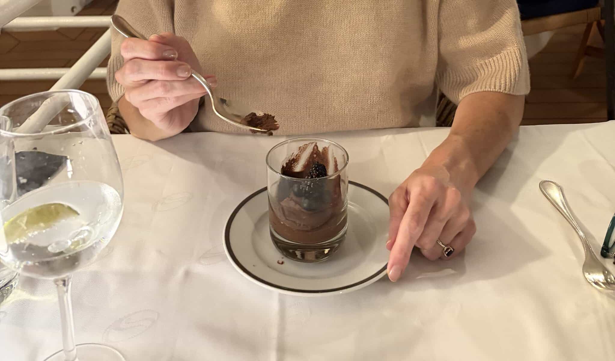Hands holding a spoon over a glass of chocolate mousse with blackberries on a white tablecloth with SeaDream logo.