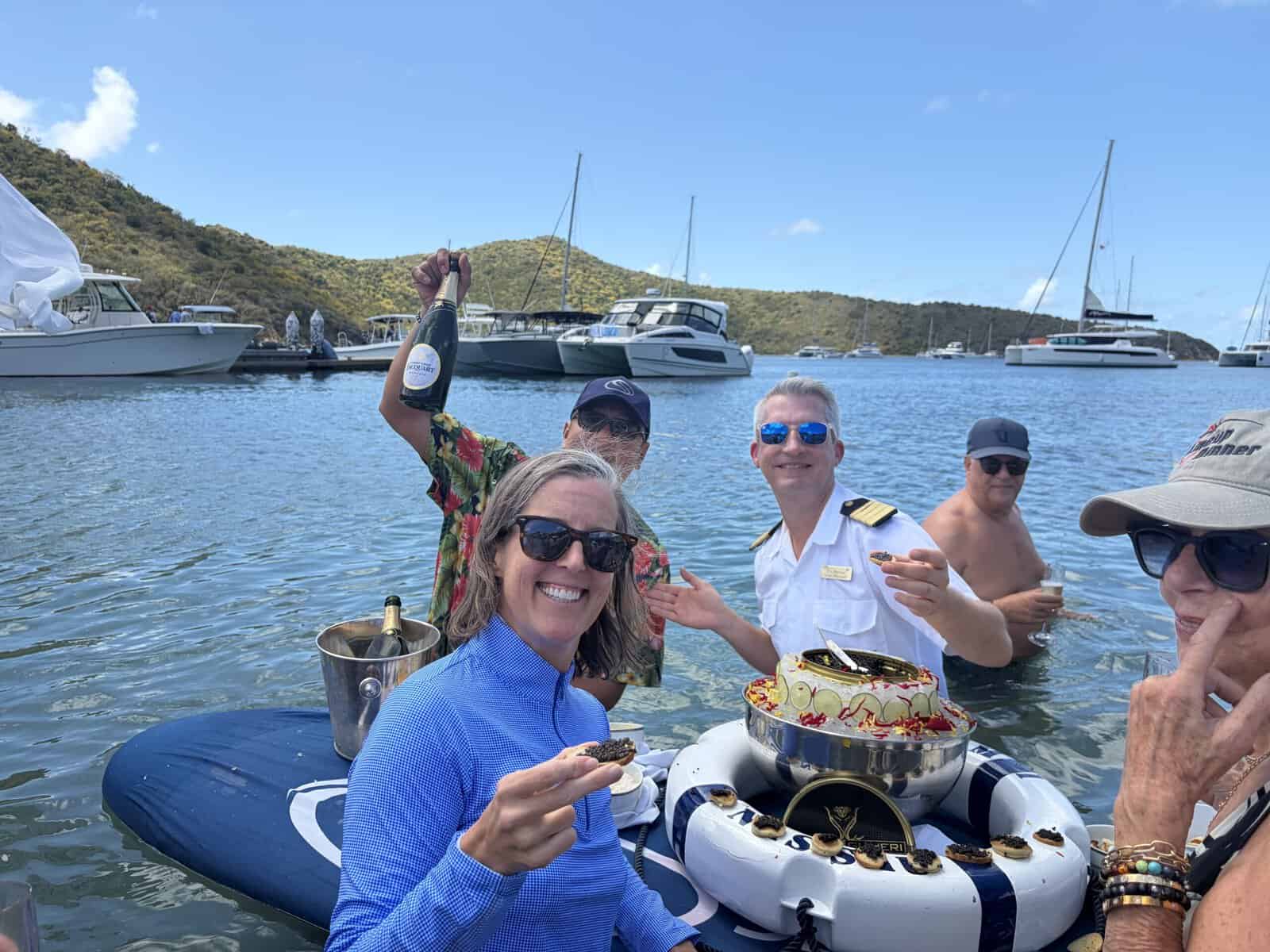 Group of guests and ship captain in shallow water enjoying caviar service and champagne with sailboats in the background.