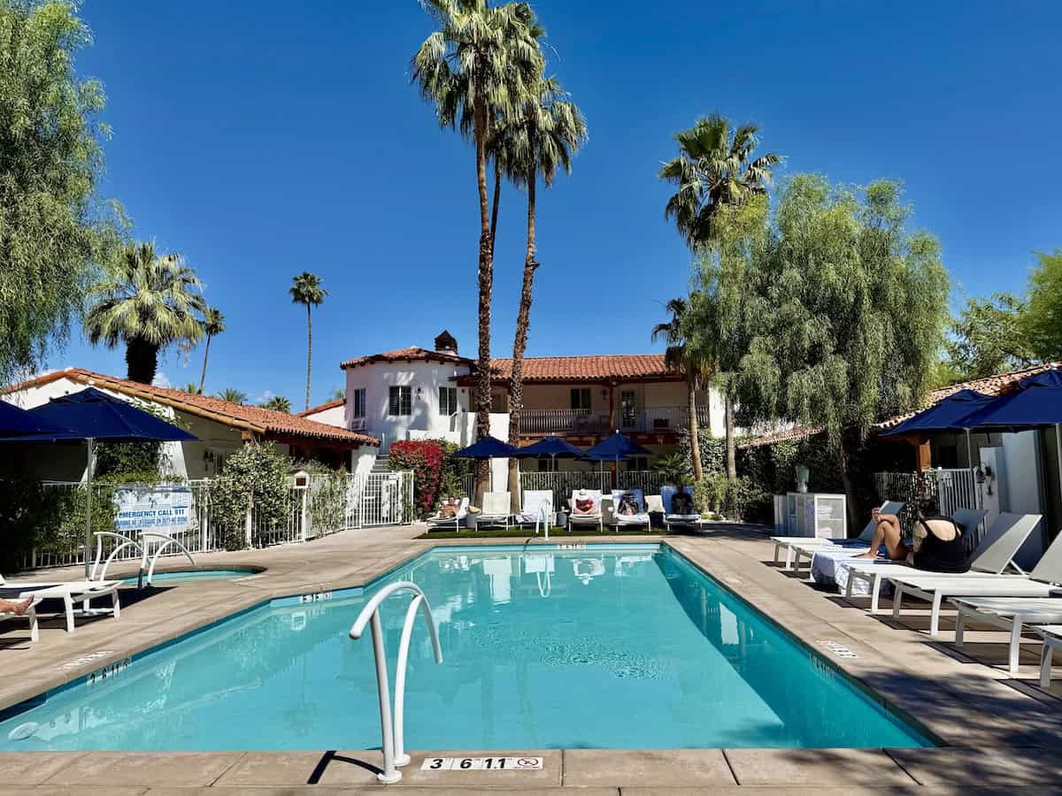 Pool at the Alcazar Palm Springs, framed by palm trees, lounge chairs, and terracotta-roofed buildings.
