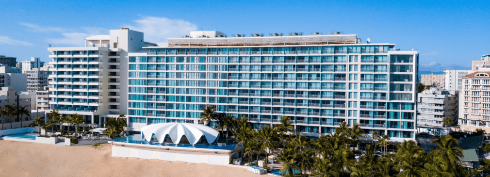 Aerial view of La Concha Resort in San Juan, Puerto Rico, showing the tall glass-fronted hotel building with rows of balconies, the white seashell-shaped Levant restaurant in front, surrounding palm trees, a sandy beach, and the Condado neighborhood in the background.
