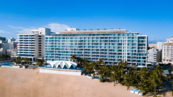 Aerial view of La Concha Resort in San Juan, Puerto Rico, showing the tall glass-fronted hotel building with rows of balconies, the white seashell-shaped Levant restaurant in front, surrounding palm trees, a sandy beach, and the Condado neighborhood in the background.