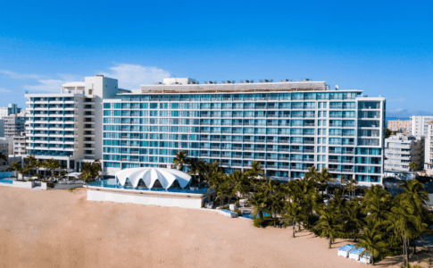 Aerial view of La Concha Resort in San Juan, Puerto Rico, showing the tall glass-fronted hotel building with rows of balconies, the white seashell-shaped Levant restaurant in front, surrounding palm trees, a sandy beach, and the Condado neighborhood in the background.