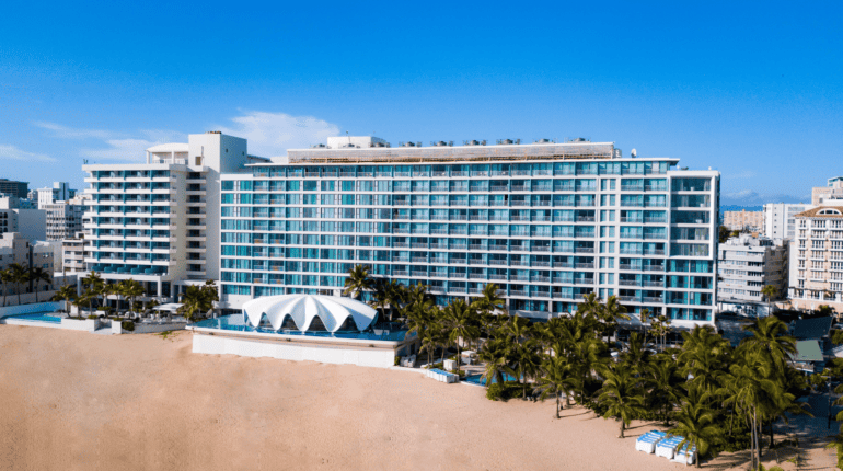 Aerial view of La Concha Resort in San Juan, Puerto Rico, showing the tall glass-fronted hotel building with rows of balconies, the white seashell-shaped Levant restaurant in front, surrounding palm trees, a sandy beach, and the Condado neighborhood in the background.