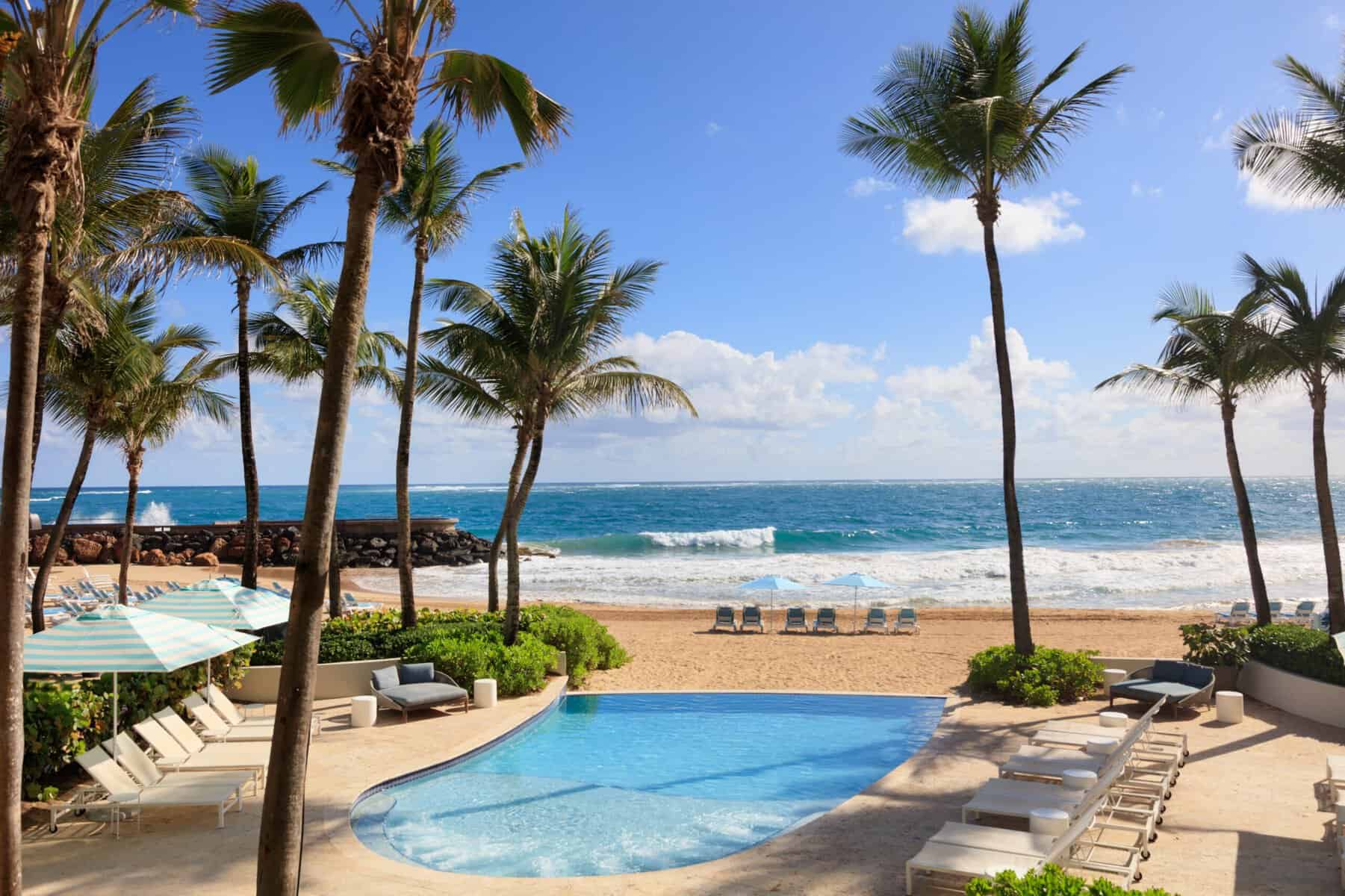 The Sereno Pool at La Concha Resort, a curved blue pool surrounded by white lounge chairs, striped teal and white umbrellas, and palm trees, with a sandy beach and the Atlantic Ocean in the background.