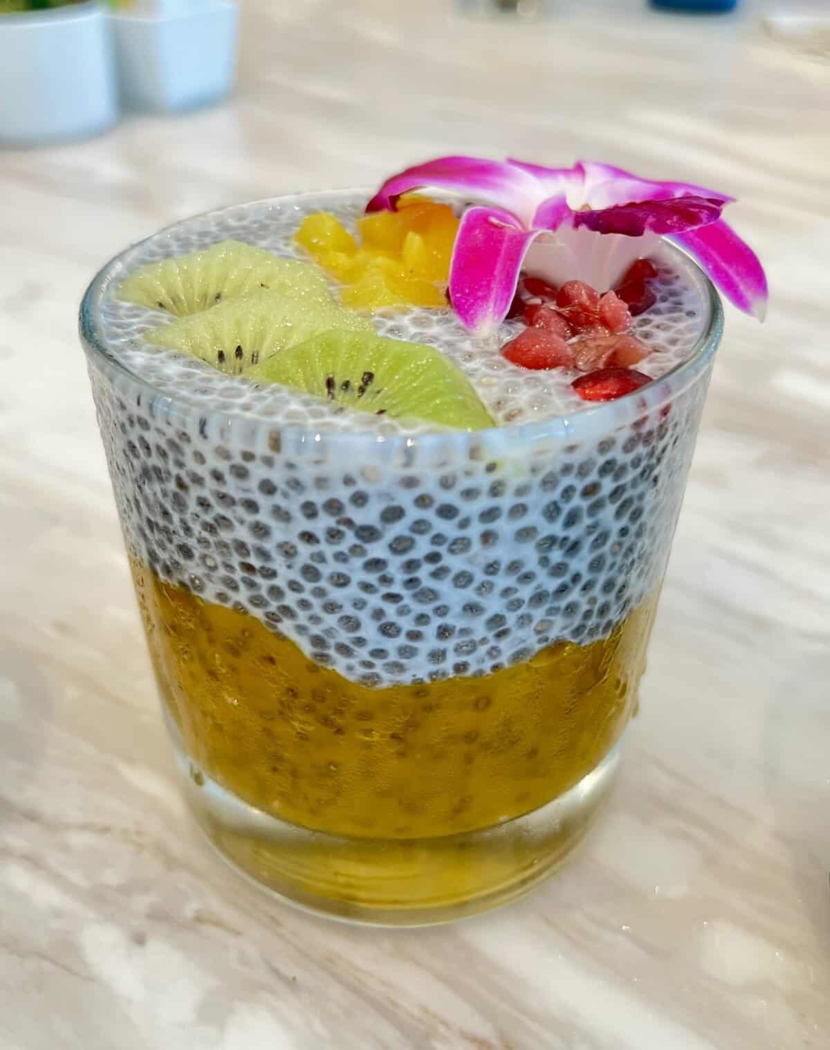 A clear glass cup holding a layered mango and chia pudding, with bright yellow mango on the bottom, white almond milk chia pudding on top, garnished with sliced kiwi, diced pineapple, pomegranate seeds, and a pink orchid flower.