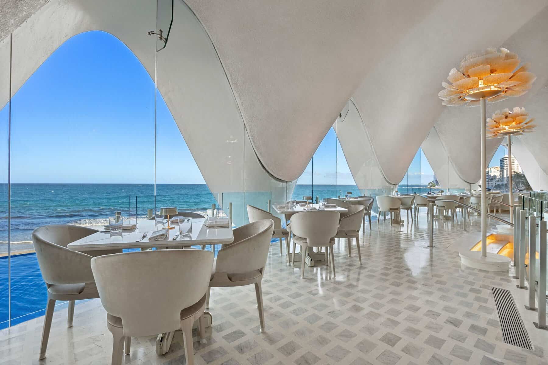 Interior of Levant restaurant at La Concha Resort, with curved white scalloped ceilings forming arched windows that overlook the Atlantic Ocean, white-and-gray patterned tile floors, cream-colored chairs around small marble tables, and flower-shaped chandeliers hanging from the ceiling.