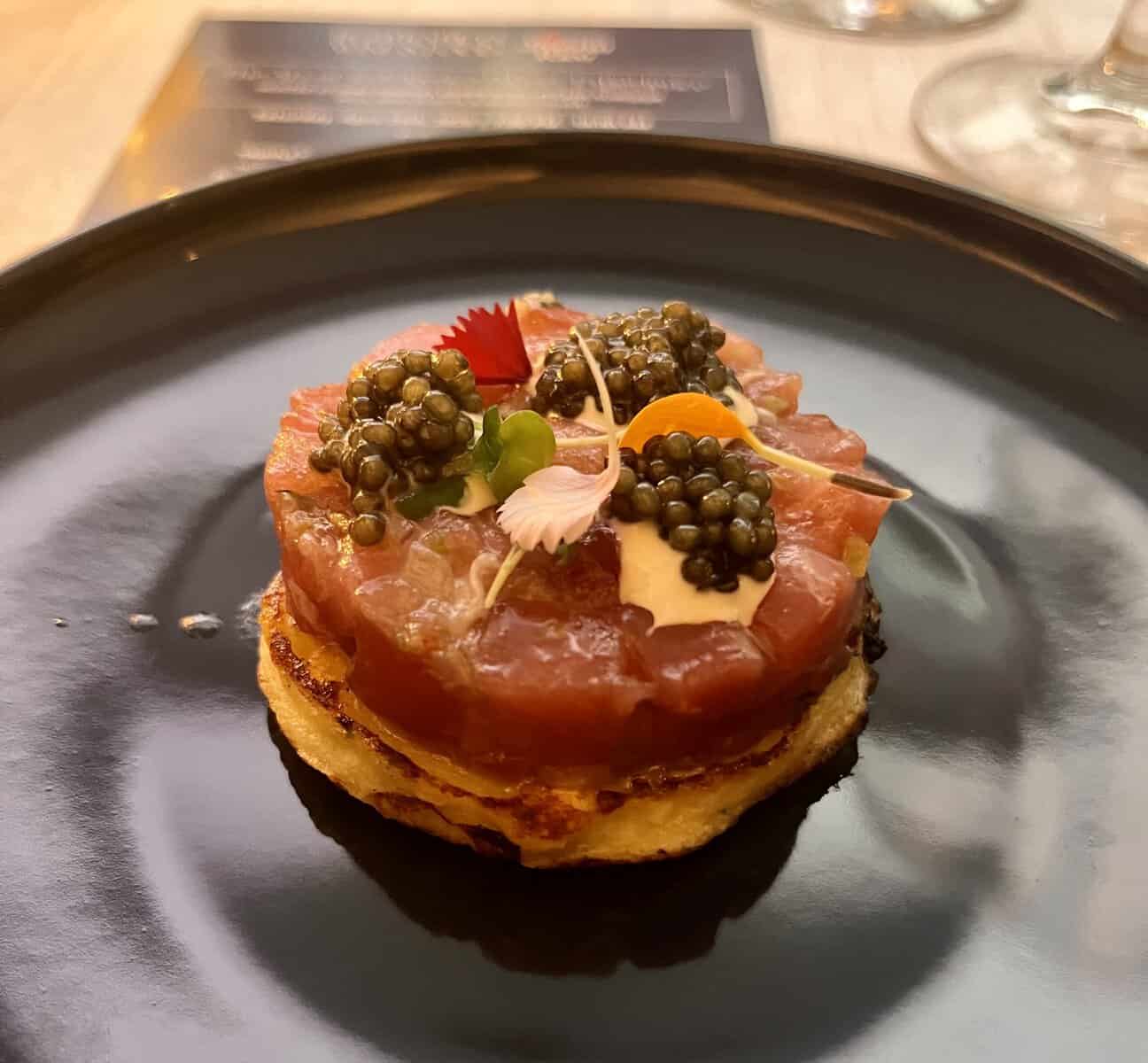 A round codfish yuca blini topped with diced bluefin tuna, three mounds of black caviar, a drizzle of coconut sofrito, and a garnish of microgreens and edible flowers, served on a black plate.