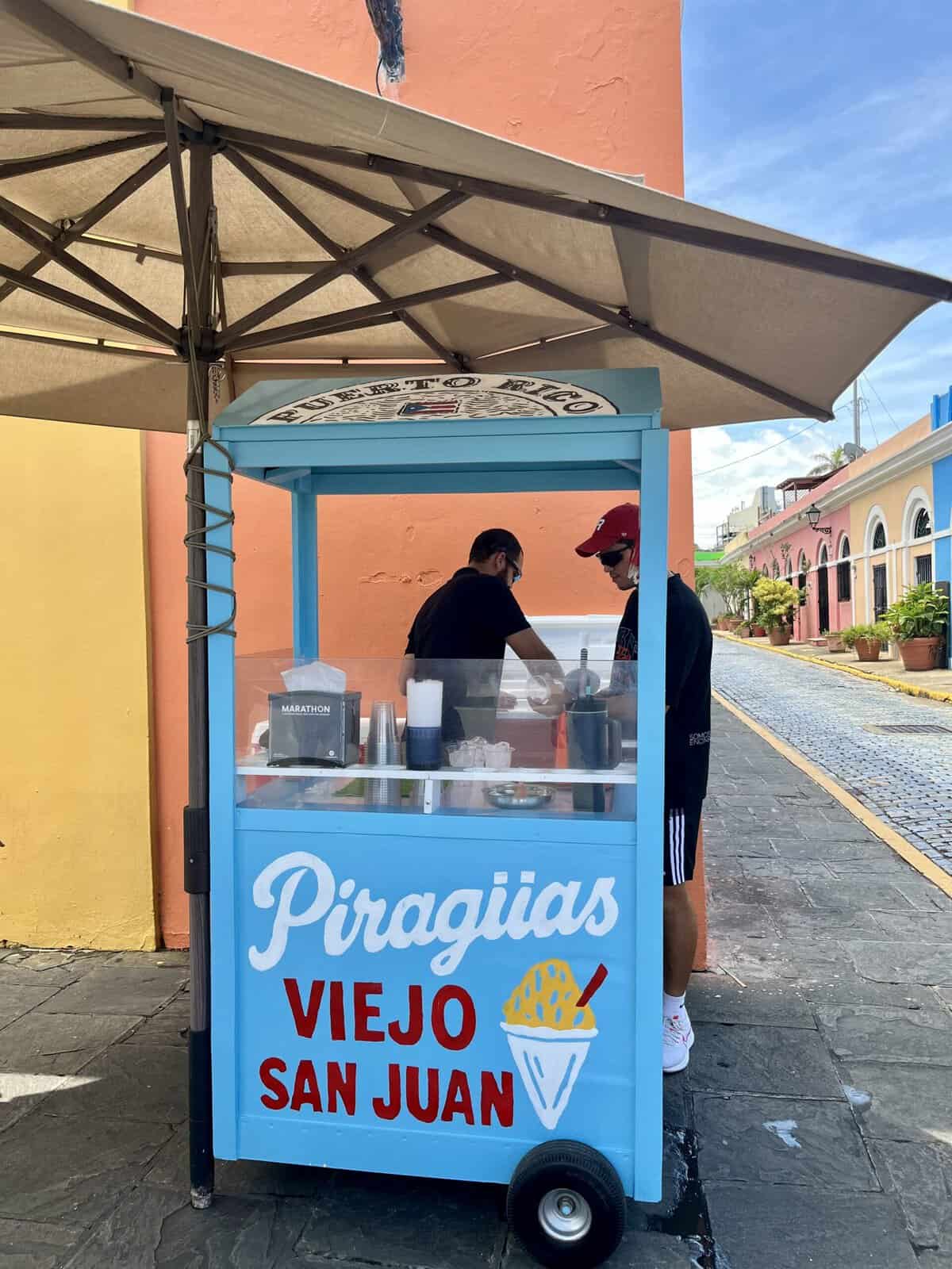 A light blue piragua cart with "Piraguas Viejo San Juan" painted on the front sits on a cobblestone street in Old San Juan, shaded by a large beige umbrella. Two men stand at the cart preparing shaved ice, with colorful orange, yellow, and pink buildings lining the street behind them.