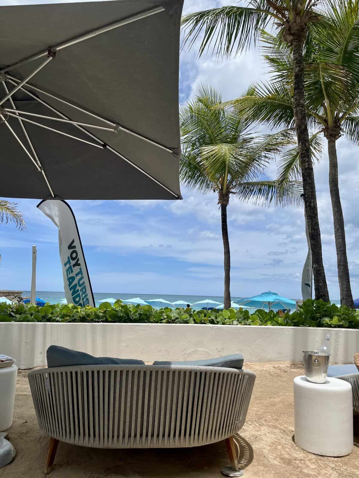 A curved gray rope daybed sits on sand beneath a large umbrella at La Concha Resort's Sereno Pool, with palm trees, blue beach umbrellas, and the Atlantic ocean visible in the background.