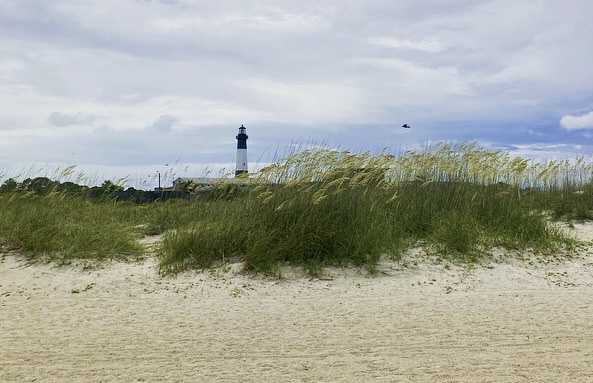 The black-and-white Tybee Island Lighthouse rises above tall dune grasses and a sandy beach in the foreground, under a cloudy gray sky.