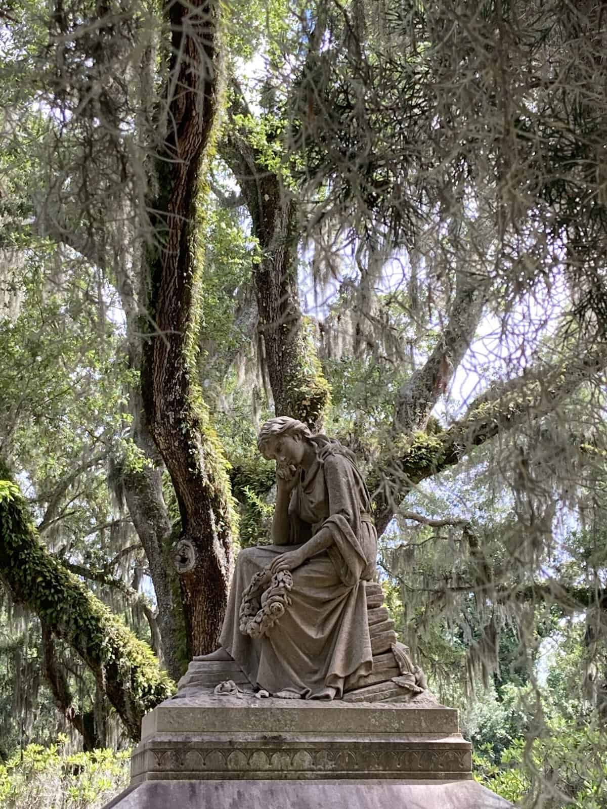 A weathered stone statue of a seated woman in flowing robes holding a wreath of flowers sits atop a pedestal at Bonaventure Cemetery, surrounded by moss-covered oak trees with Spanish moss hanging from the branches.