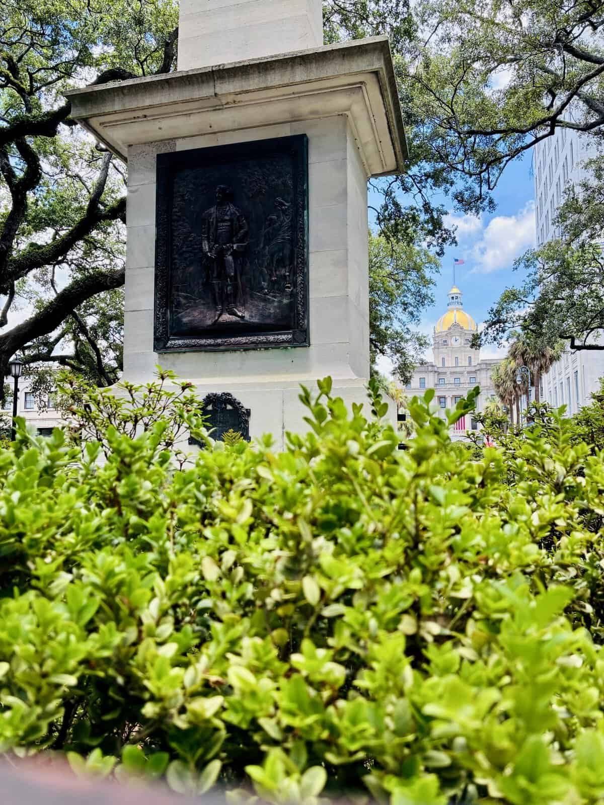 A stone monument with a bronze relief plaque depicting a historical figure rises above a hedge of green shrubs in Johnson Square, Savannah. In the background, the gold-domed Savannah City Hall stands under a blue sky with an American flag flying on top.