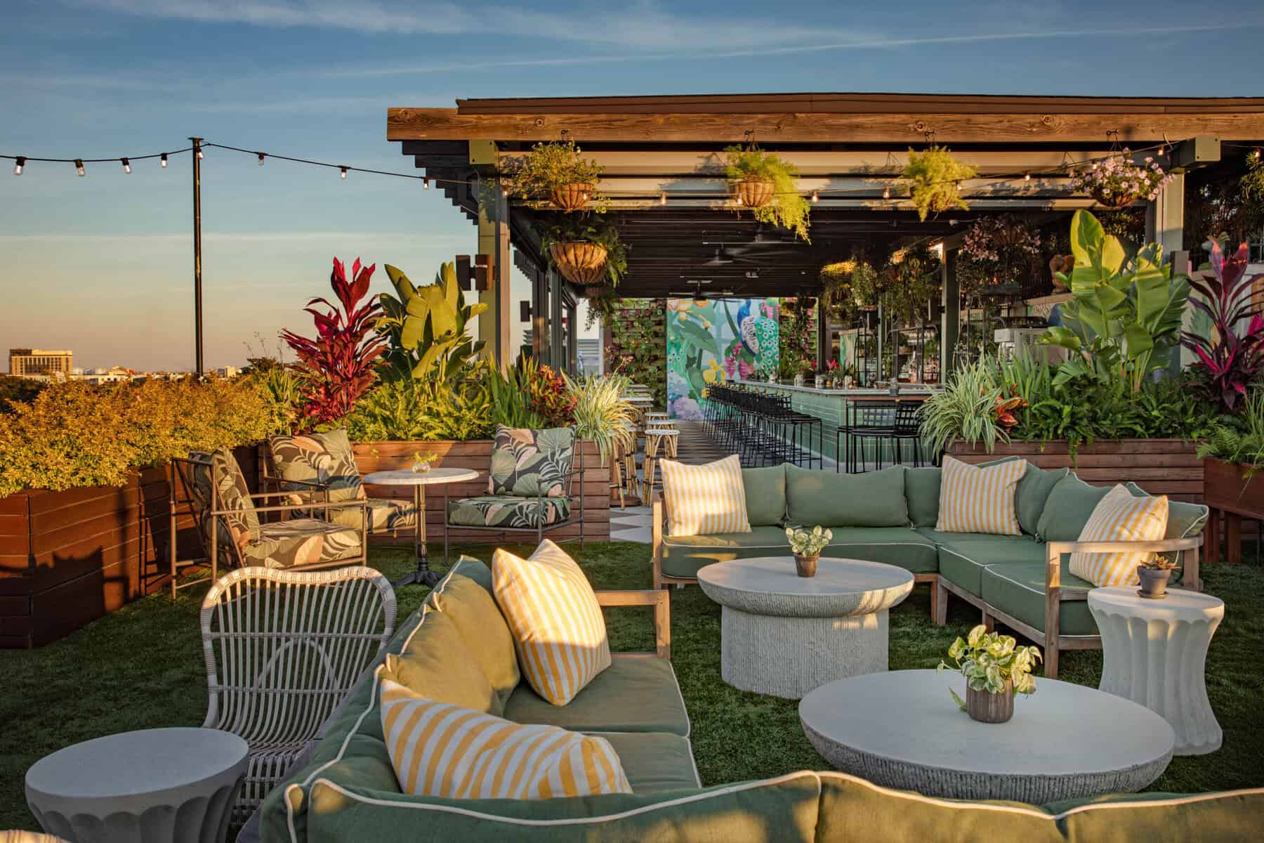 The rooftop bar and lounge area at Perry Lane Hotel, with green sectional sofas topped with yellow and white striped pillows, wicker chairs, round concrete coffee tables on artificial turf, and wooden planters filled with tropical foliage. A bar with black stools sits under a wooden pergola with hanging ferns and string lights, with a colorful floral mural in the background and the Savannah skyline visible at sunset.