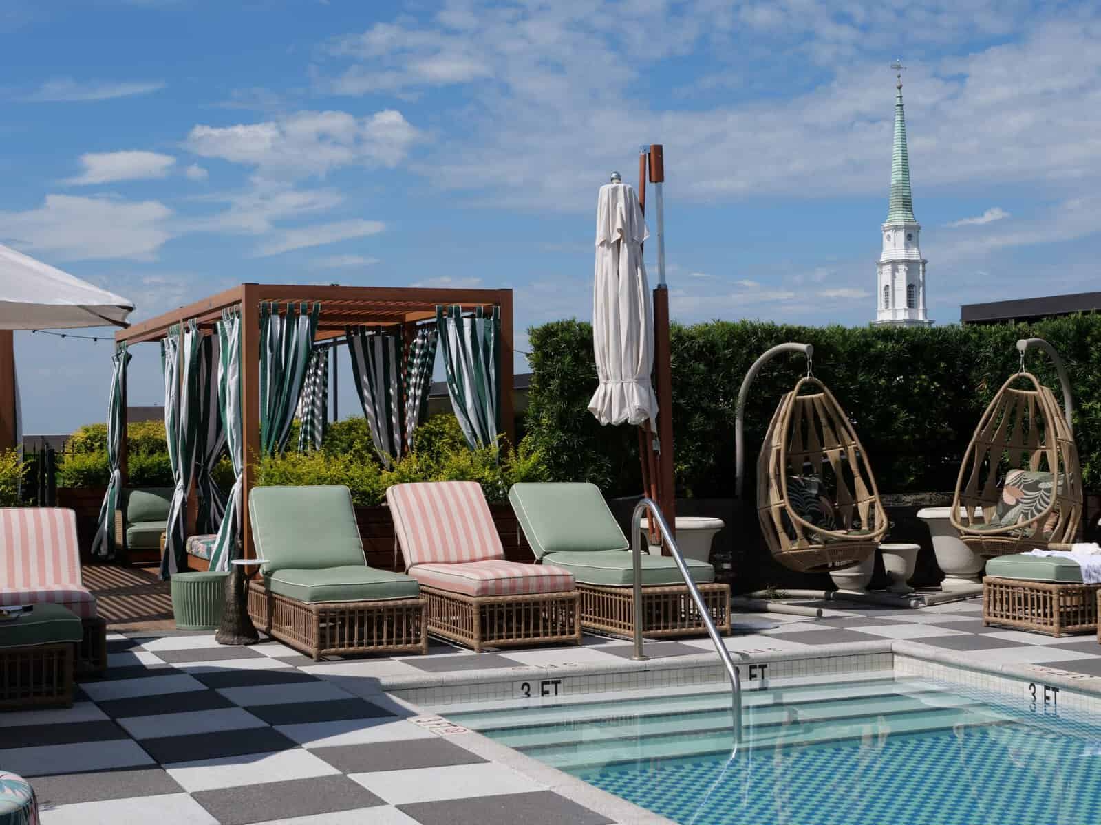 The rooftop pool at Perry Lane Hotel, with a checkered black-and-white tile deck, wicker loungers cushioned in sage green and pink stripes, hanging rattan egg chairs, and a green-and-white striped cabana. A white church steeple rises above the treeline in the background under a blue sky.