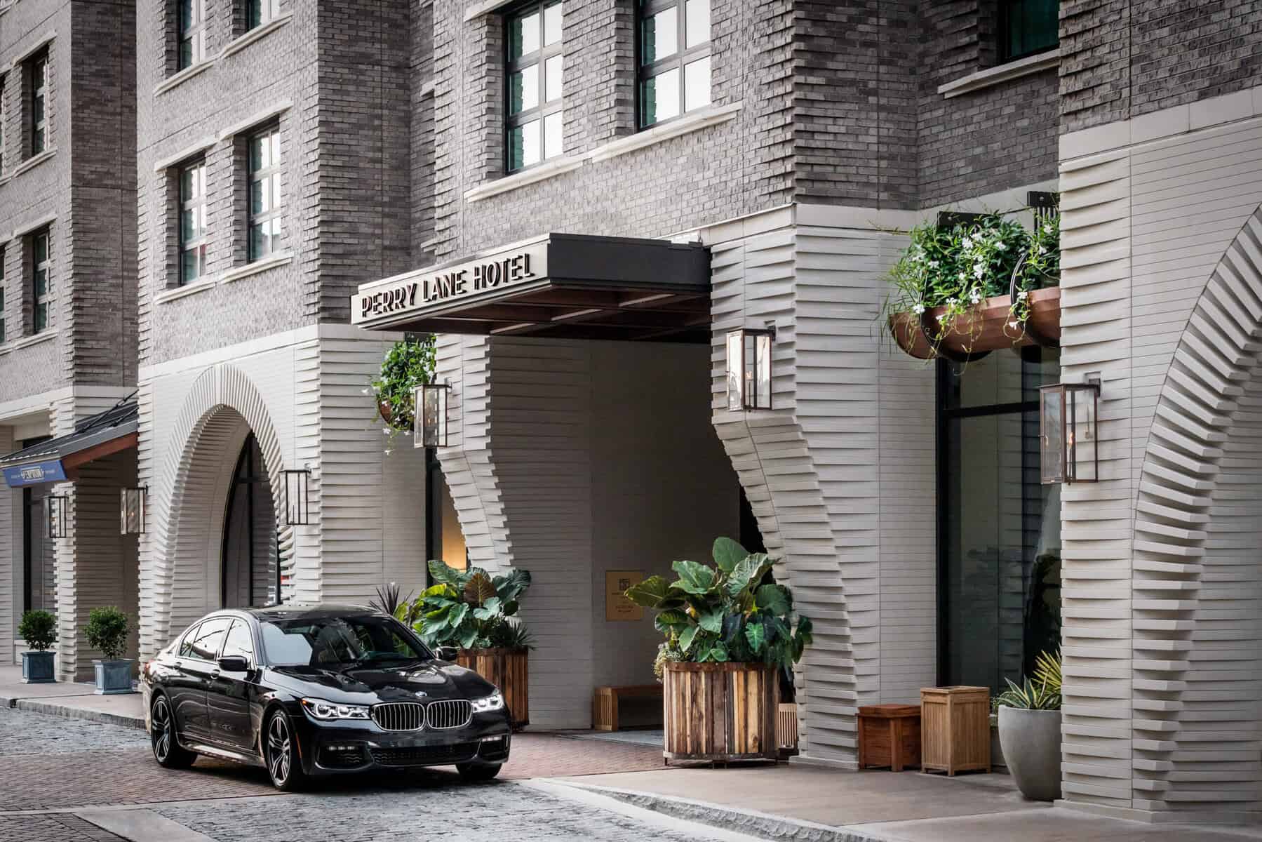 The exterior entrance of Perry Lane Hotel, featuring a gray brick facade with white ribbed archways, hanging lanterns, planters filled with tropical greenery, and a black BMW parked out front on a cobblestone street.