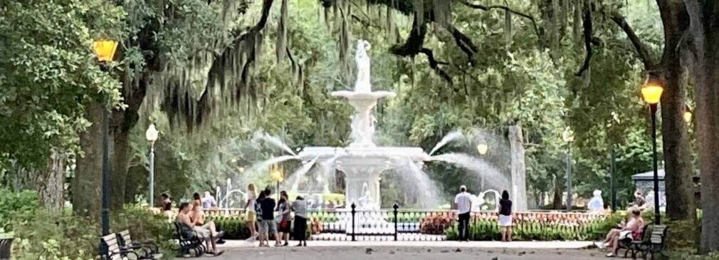 A wide walkway lined with benches leads toward the white tiered Forsyth Park fountain, with water sprays visible around it. Massive oak trees draped in Spanish moss arch overhead, and visitors sit on benches and stand near the fountain.