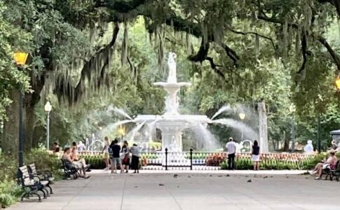 A wide walkway lined with benches leads toward the white tiered Forsyth Park fountain, with water sprays visible around it. Massive oak trees draped in Spanish moss arch overhead, and visitors sit on benches and stand near the fountain.