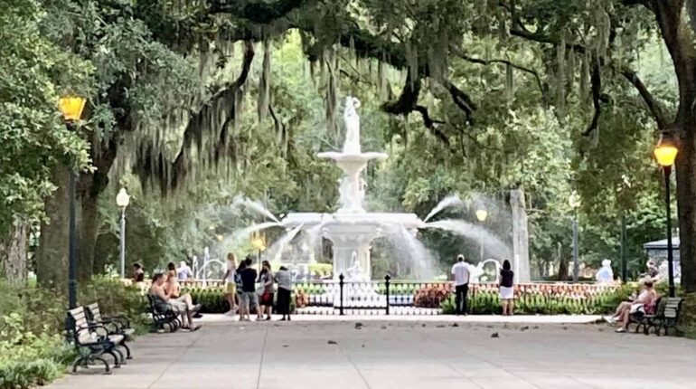 A wide walkway lined with benches leads toward the white tiered Forsyth Park fountain, with water sprays visible around it. Massive oak trees draped in Spanish moss arch overhead, and visitors sit on benches and stand near the fountain.