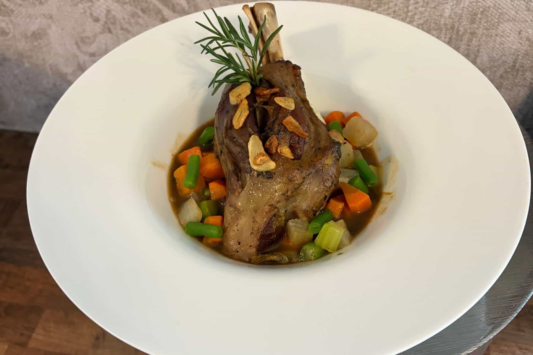 A braised lamb shank in a white bowl with diced vegetables and rosemary.