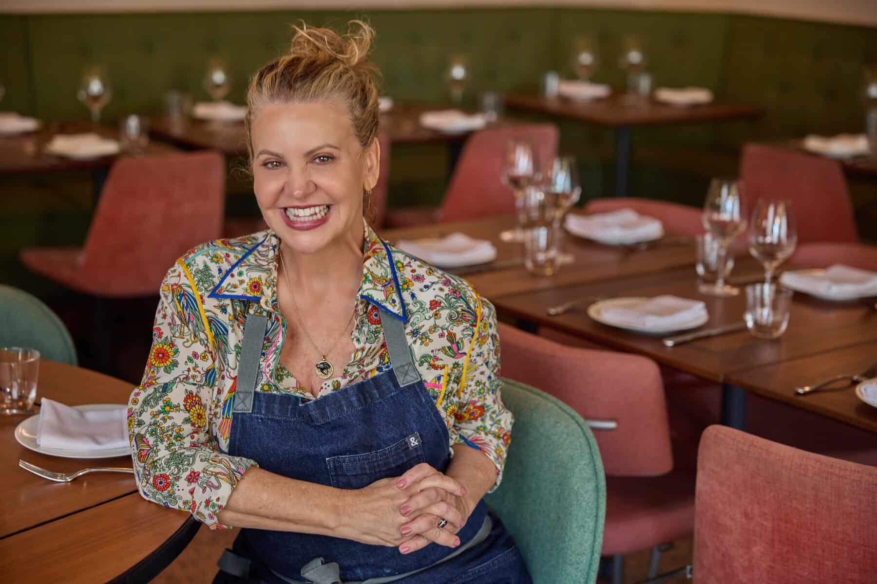 A smiling chef in a denim apron seated in a dining room.A smiling chef in a denim apron seated in a dining room. 