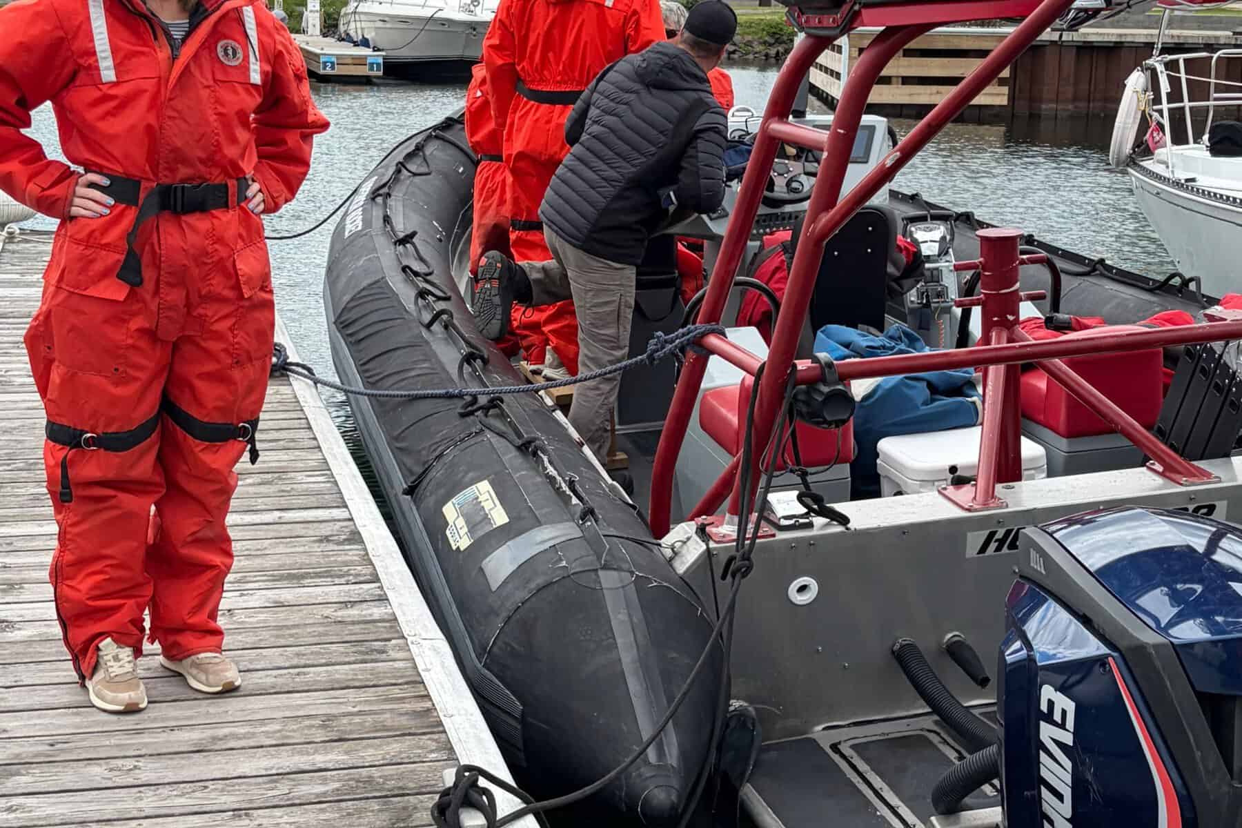 A woman in an orange flotation suit stands on a dock beside a Zodiac.