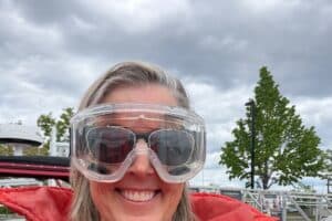 A smiling woman in an orange flotation suit and clear goggles at a marina.