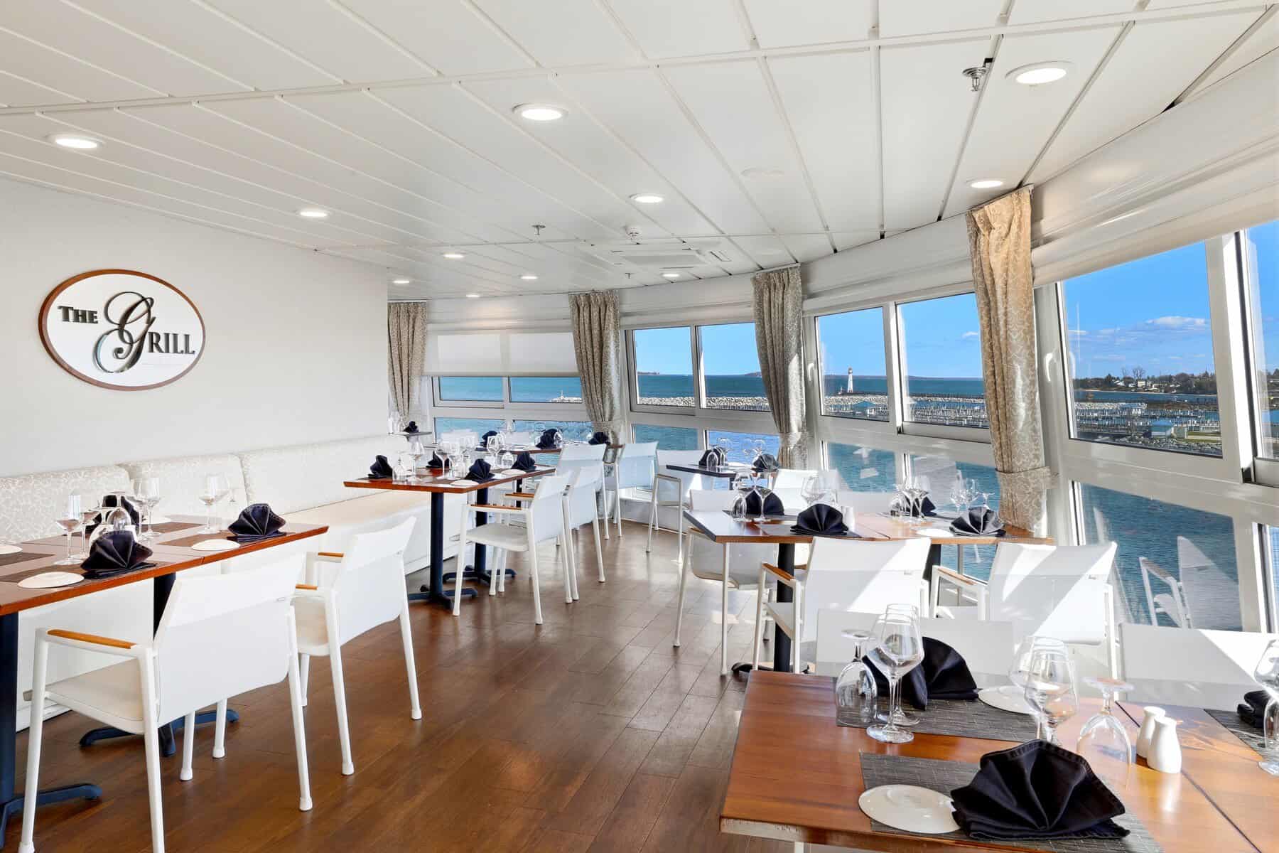 A bright shipboard restaurant with white chairs and harbor-view windows.